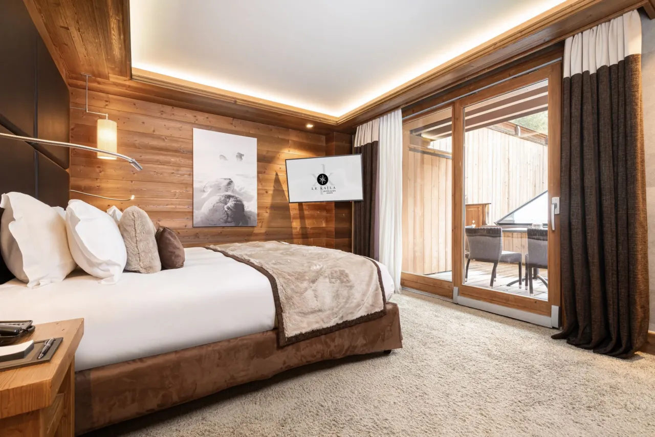Chambre aux murs en bois avec grand lit et baie vitrée donnant sur un balcon meublé.