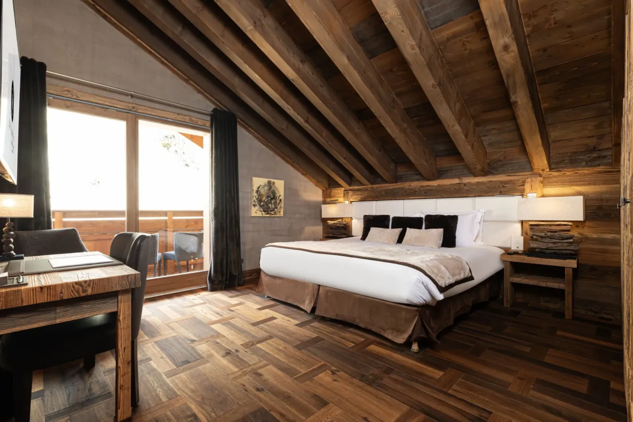 Chambre sous une charpente apparente en bois, avec lit double, bureau et baie vitrée donnant sur un balcon.