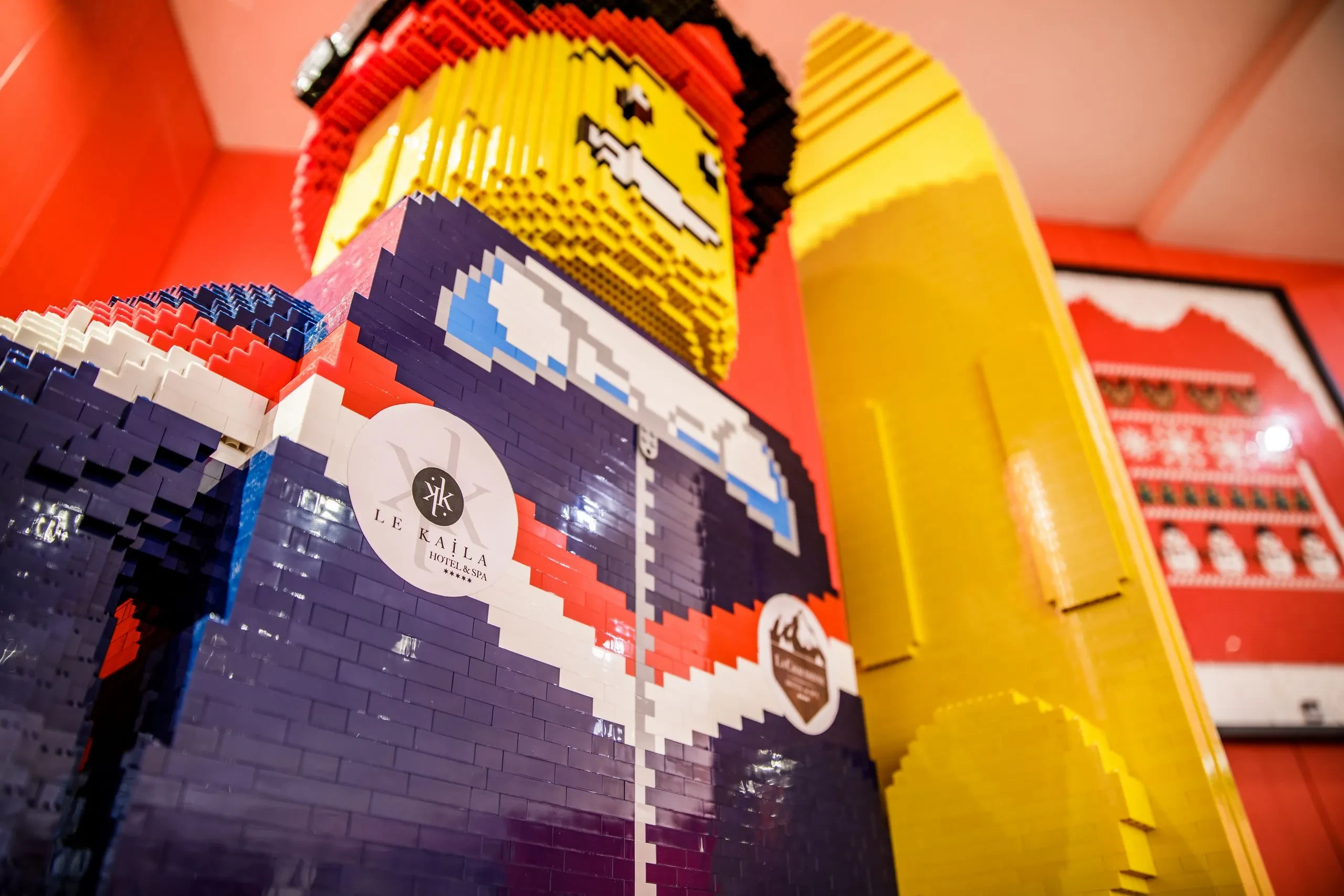 Skieur géant en briques LEGO, portant le logo Le Kaïla et tenant un ski jaune, vu en contre-plongée.