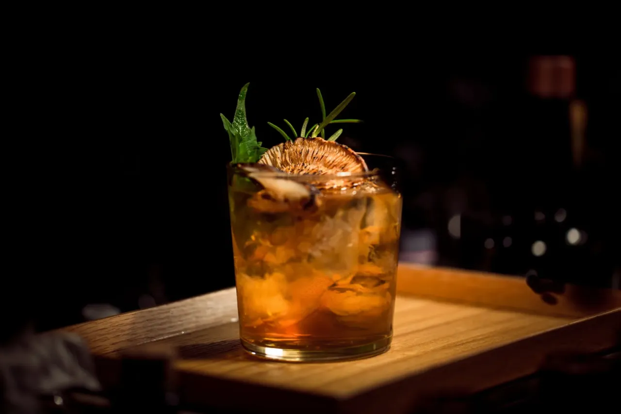 Cocktail ambré sur glaçons dans un verre bas, garni d’un agrume séché et d’herbes aromatiques.