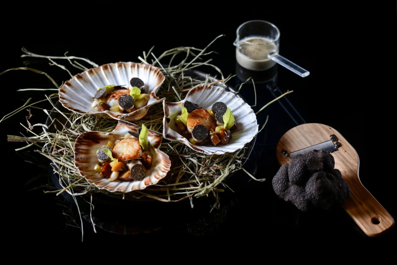 Trois noix de Saint-Jacques dorées, garnies de lamelles de truffe et servies dans leurs coquilles sur du foin.