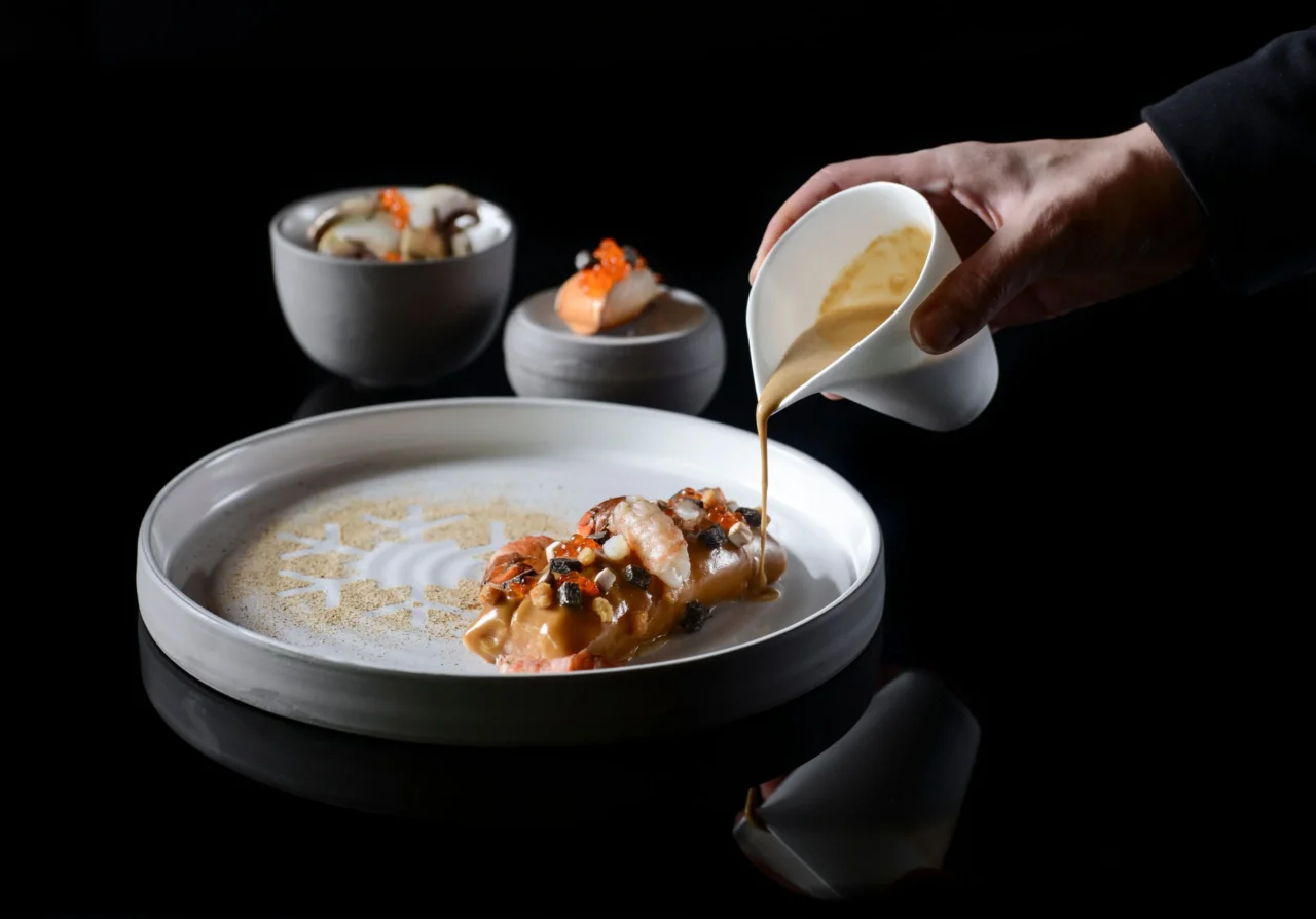 Une main verse une sauce sur un plat de crustacés garni de petites touches orangées, dans une grande assiette blanche.