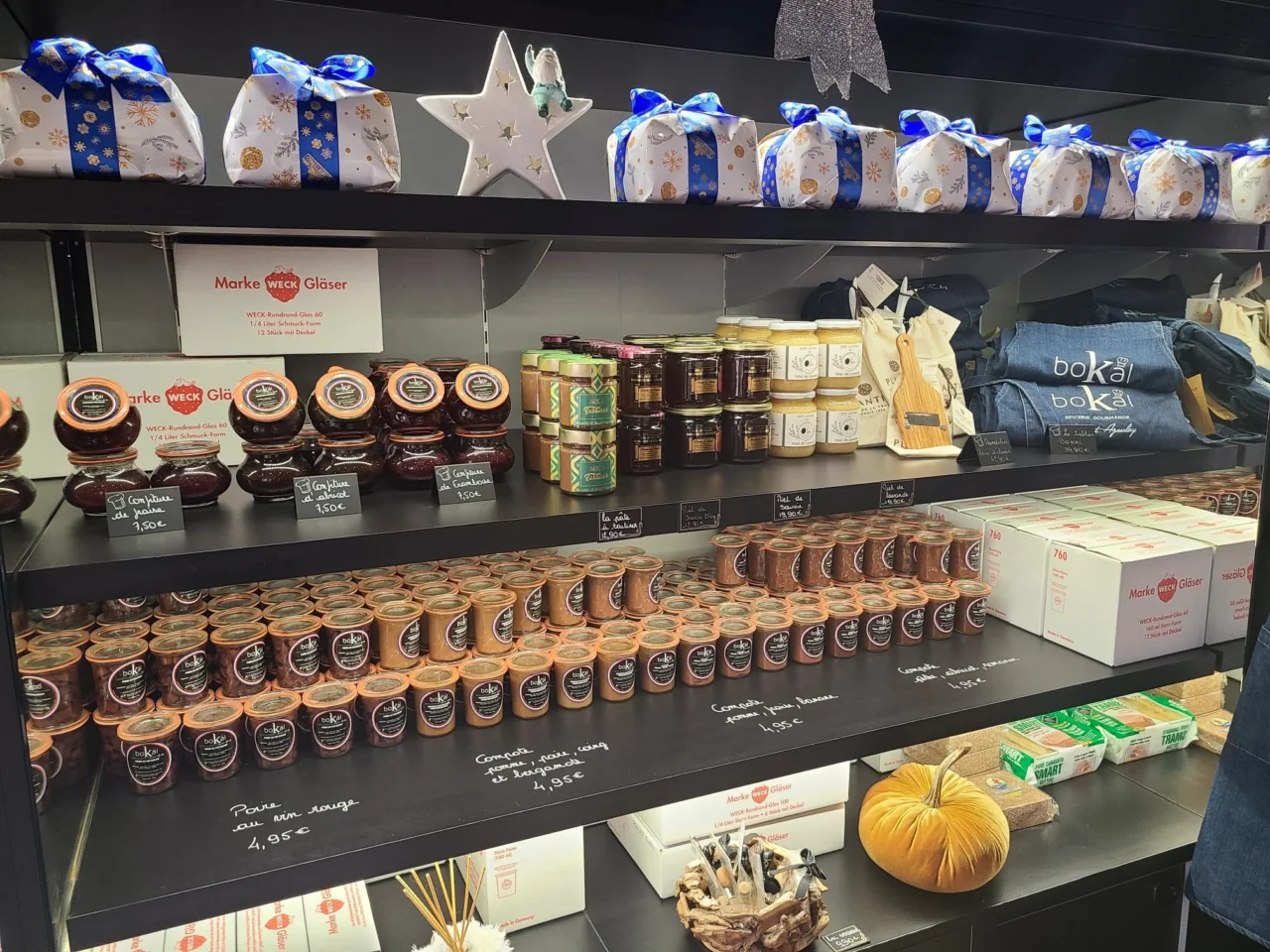 Étagères d’épicerie garnies de confitures, de miels et de préparations en bocaux, surmontées de paquets cadeaux.