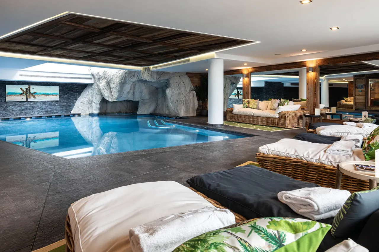 Piscine intérieure bordée de transats, avec décor de grotte et plafond en bois.