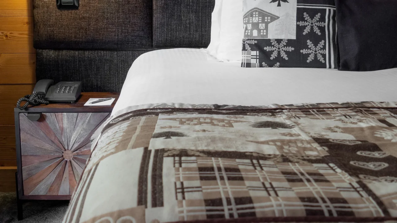 Lit de l’Hôtel La Chaudanne avec couverture et coussins à motifs montagnards, près d’un chevet en bois.