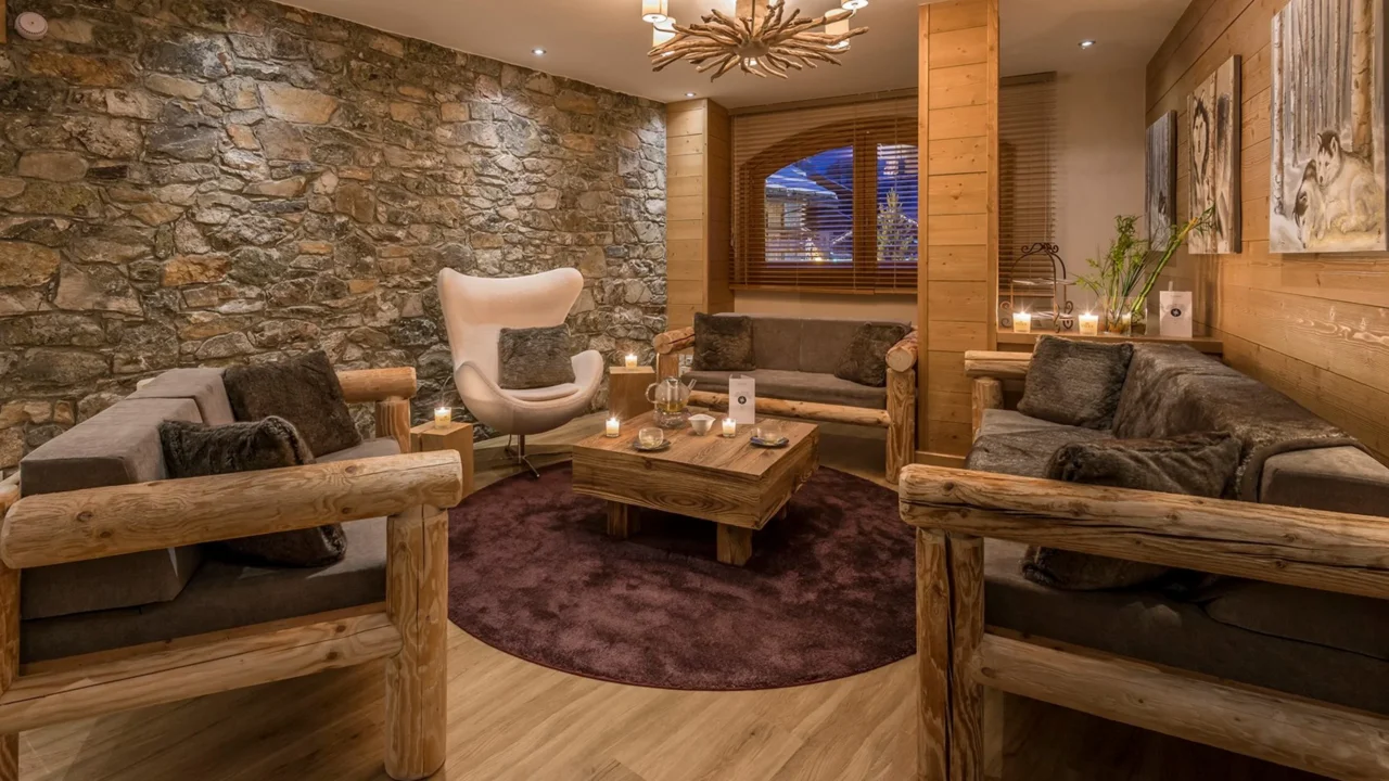 Salon de l’Hôtel La Chaudanne avec canapés en bois brut et fauteuil autour d’une table basse, près d’un mur en pierre.