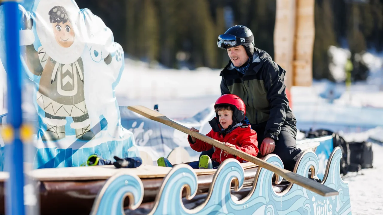 Un enfant casqué tient une rame en bois dans un bateau décoratif, avec un adulte assis derrière lui dans la neige.