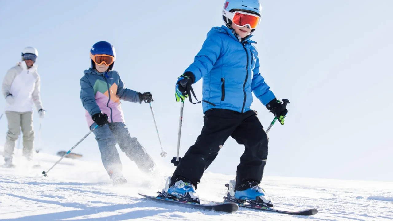 Deux enfants casqués descendent une piste de ski enneigée, suivis d’un adulte.