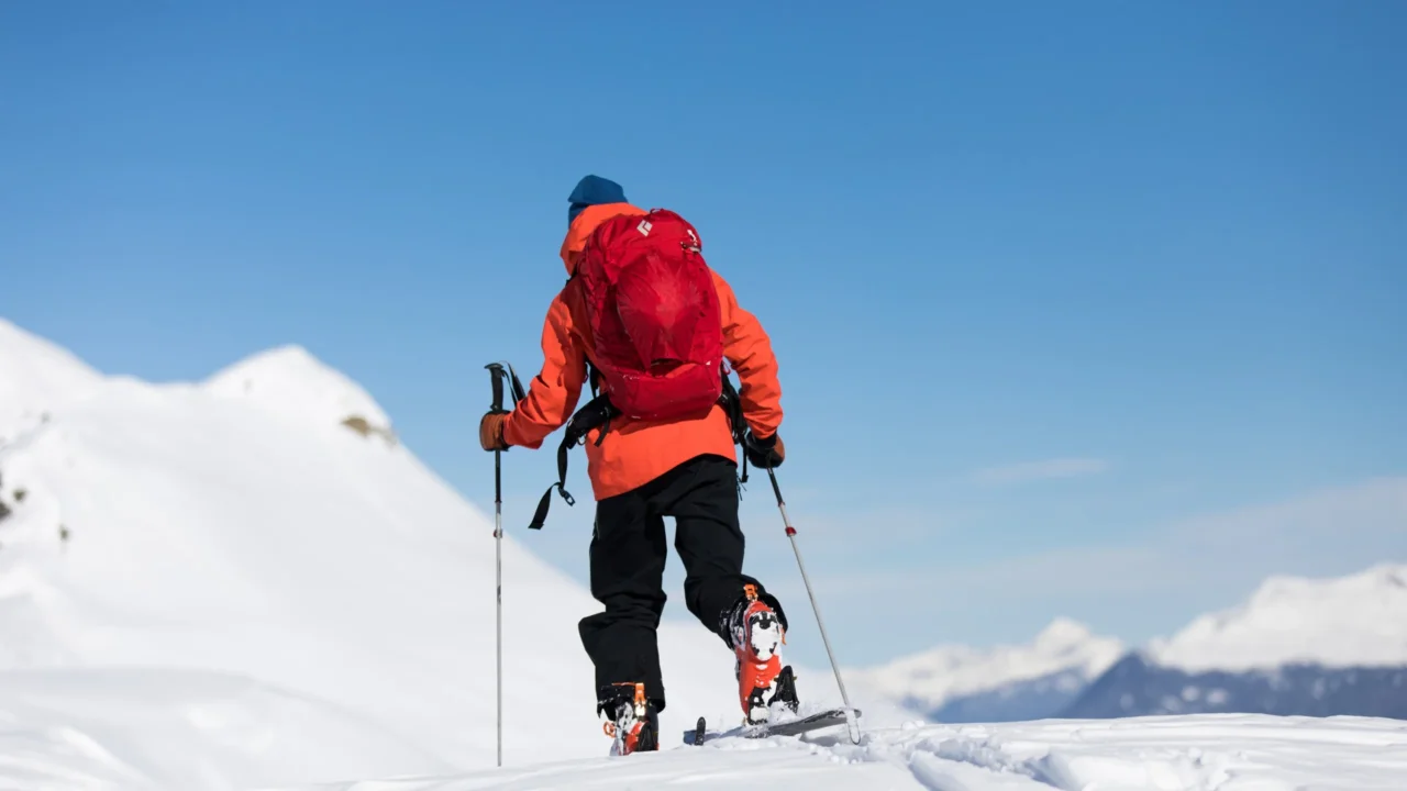 Une personne en ski de randonnée gravit une pente enneigée, avec des bâtons et un sac à dos rouge.