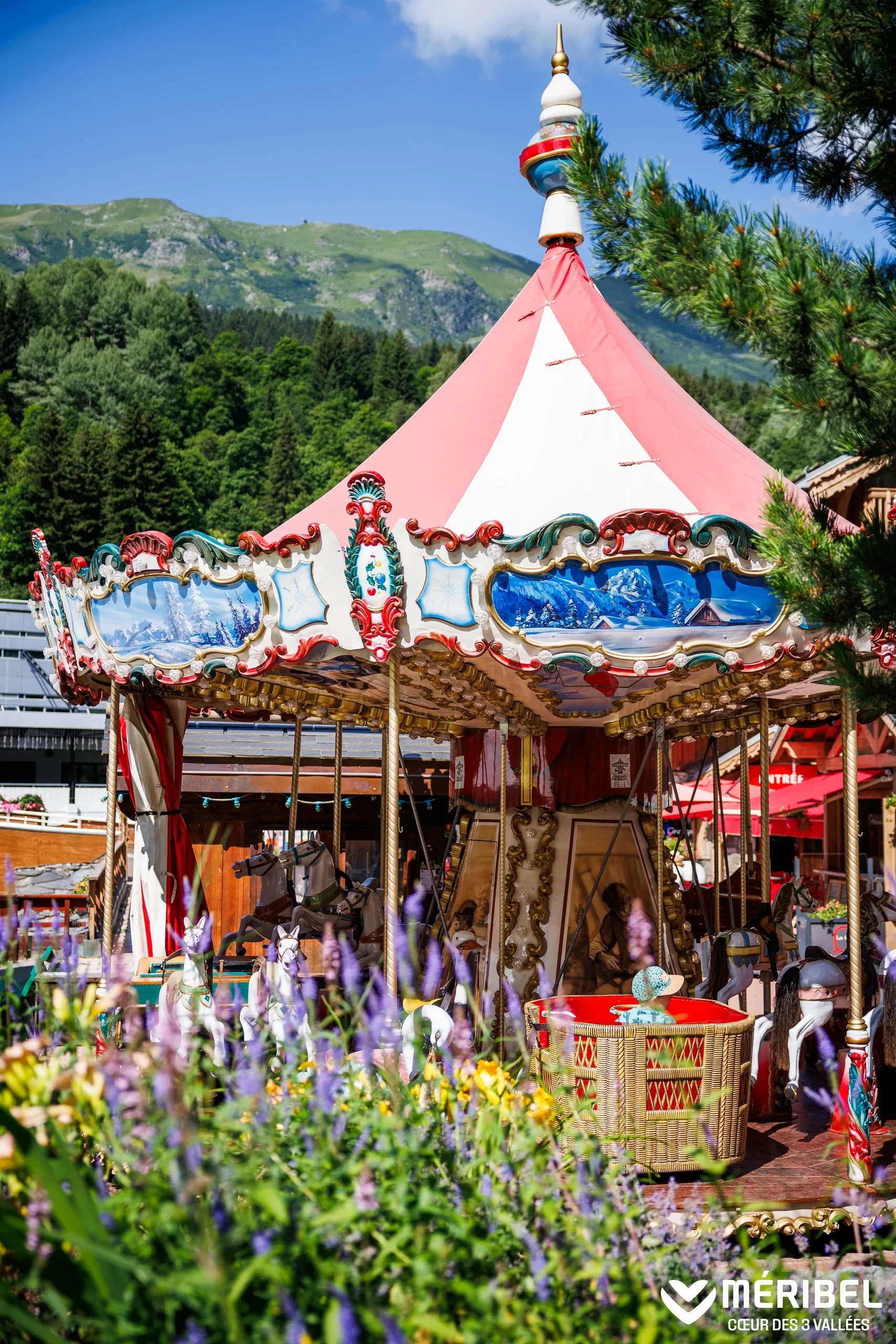 Manège à chevaux de bois à Méribel, entouré de fleurs, avec les montagnes en arrière-plan.