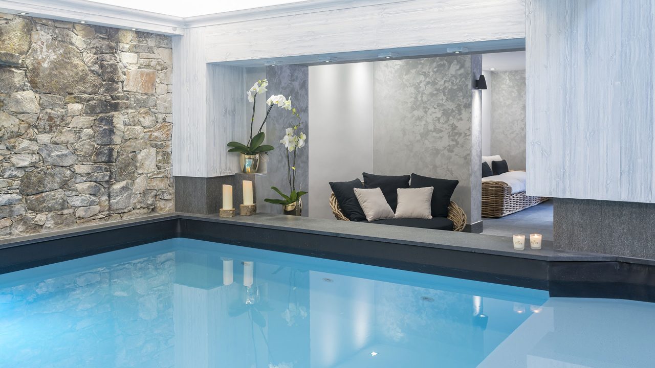Piscine intérieure bordée de pierre, avec une banquette à coussins et des transats dans l’espace de repos adjacent.