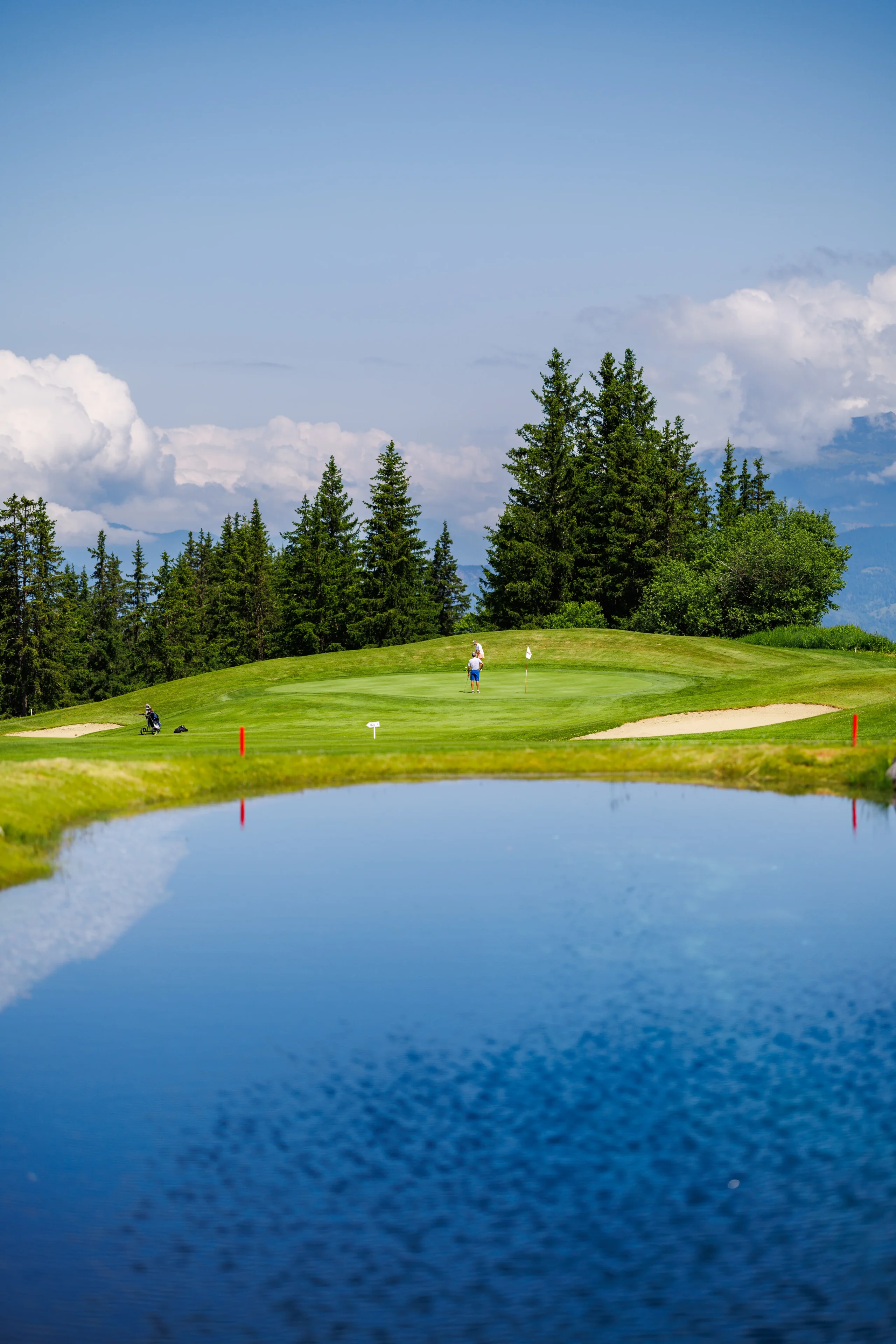 Deux golfeurs sur un green bordé de bunkers et de conifères, derrière un plan d’eau.