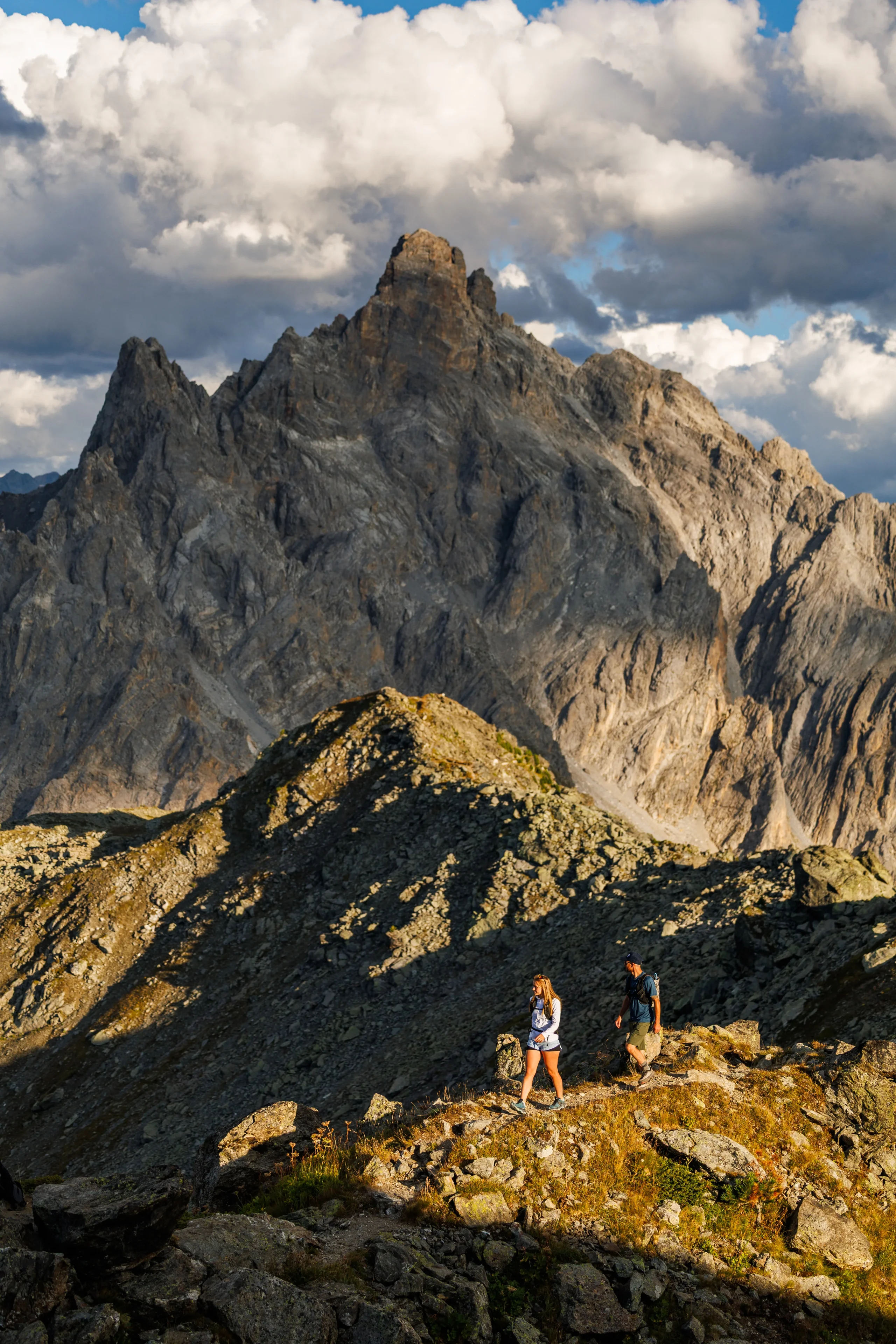 Deux randonneurs sur un sentier rocheux au pied de hautes aiguilles montagneuses.
