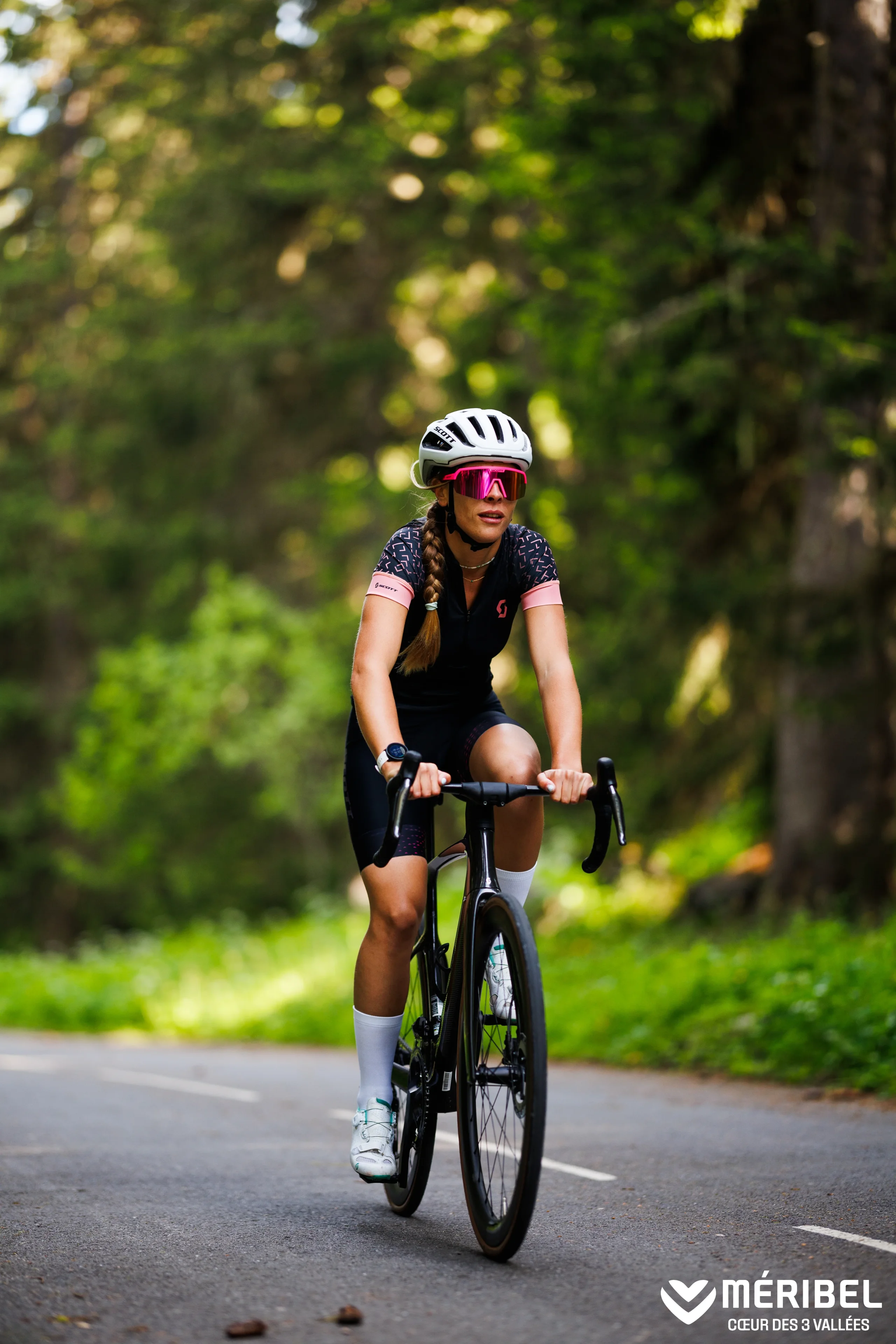 Cycliste portant un casque blanc et des lunettes roses sur un vélo de route, sur une route en forêt à Méribel.