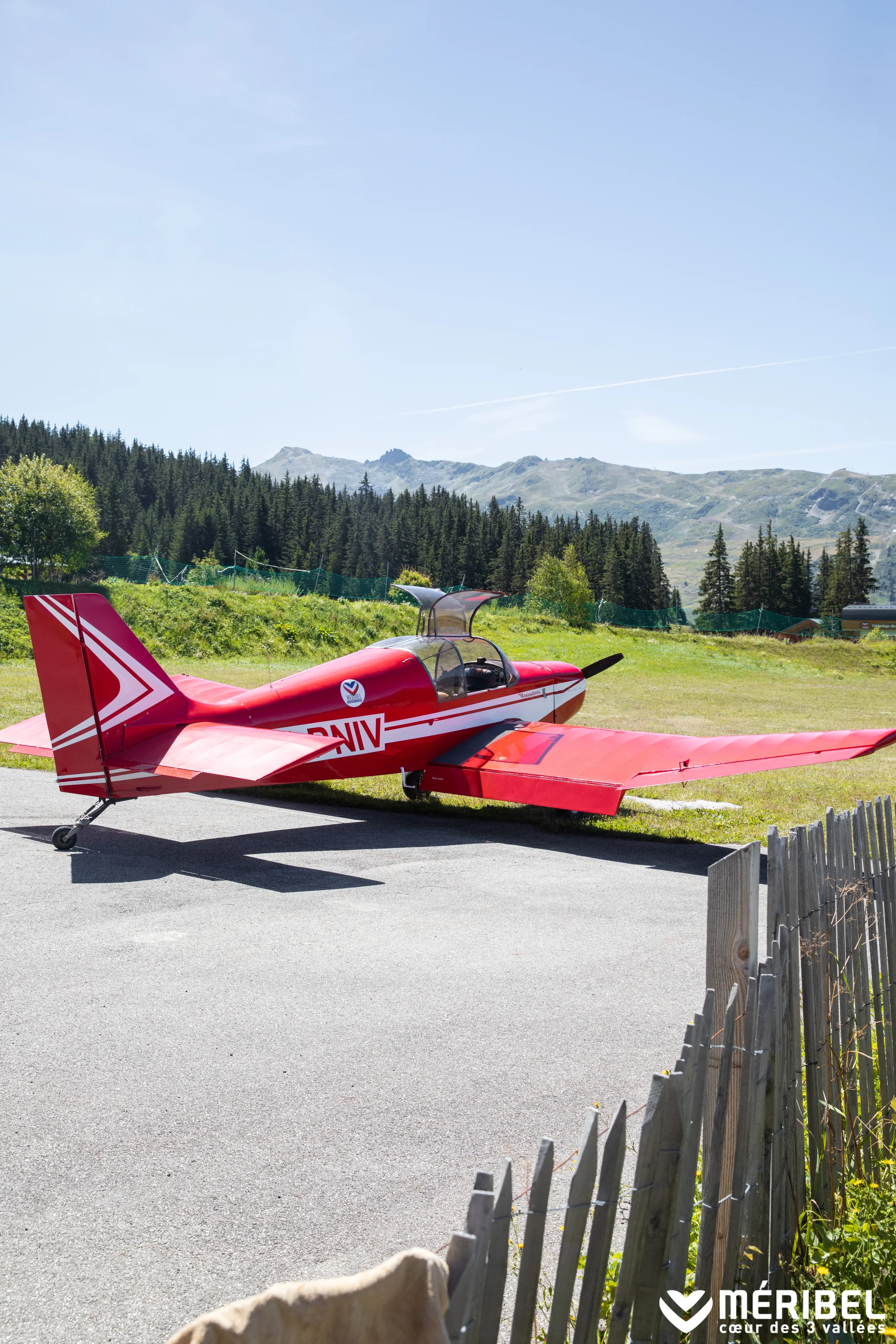 Avion de tourisme rouge et blanc stationné devant une forêt et des reliefs montagneux à Méribel.