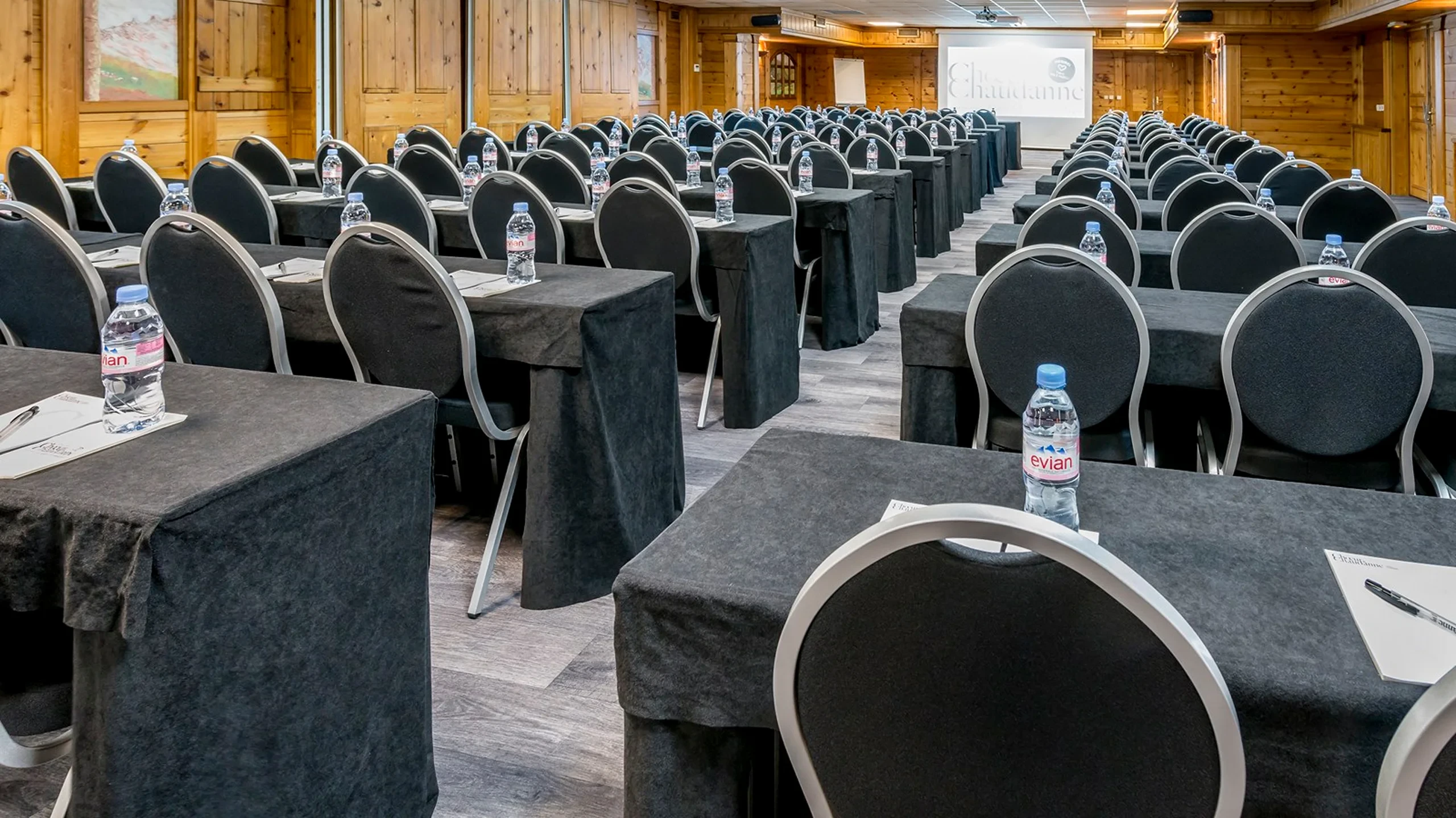 Salle boisée aménagée avec des rangées de tables et de chaises face à un écran de projection, de part et d’autre d’une allée centrale.
