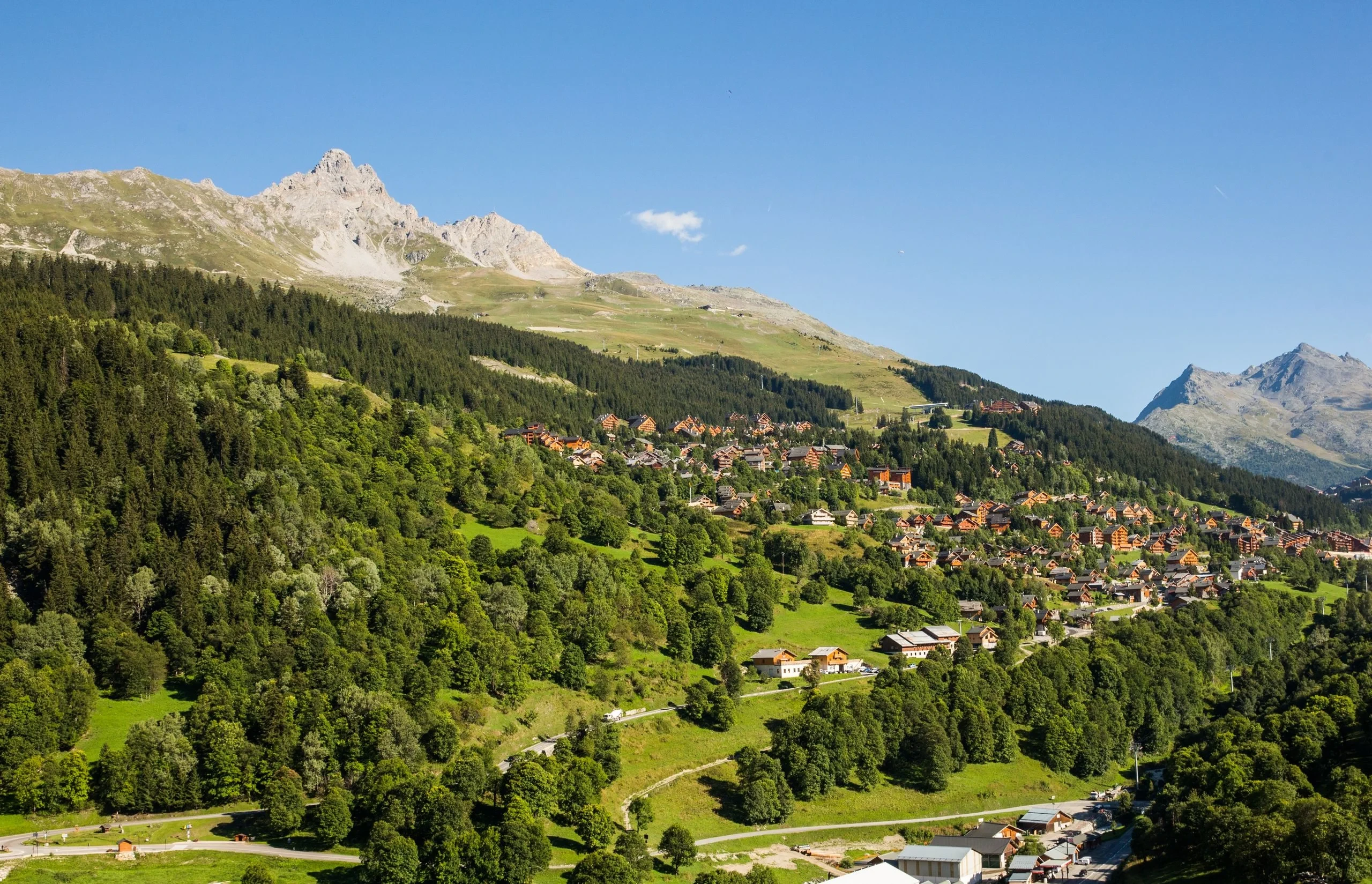 Village de chalets sur un versant verdoyant, entre forêts et sommets rocheux.