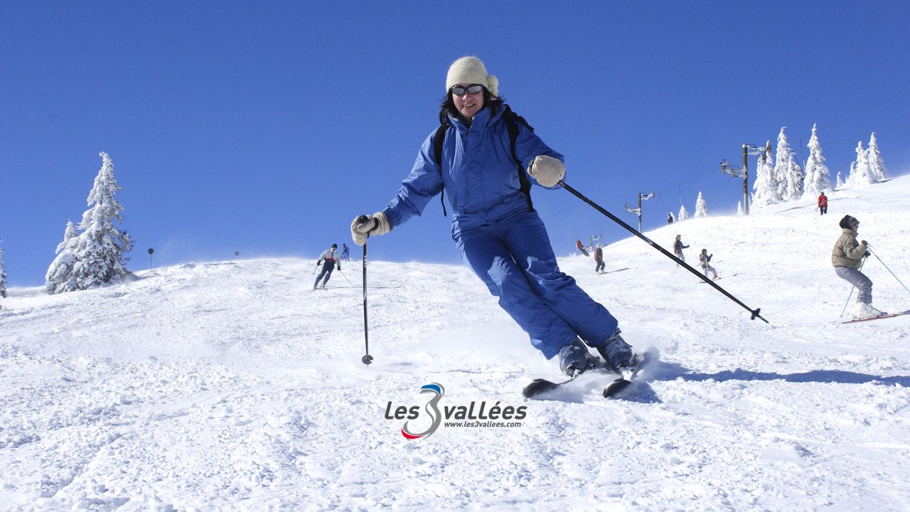 Skieuse en tenue bleue prenant un virage sur une piste enneigée, avec d’autres skieurs en arrière-plan.