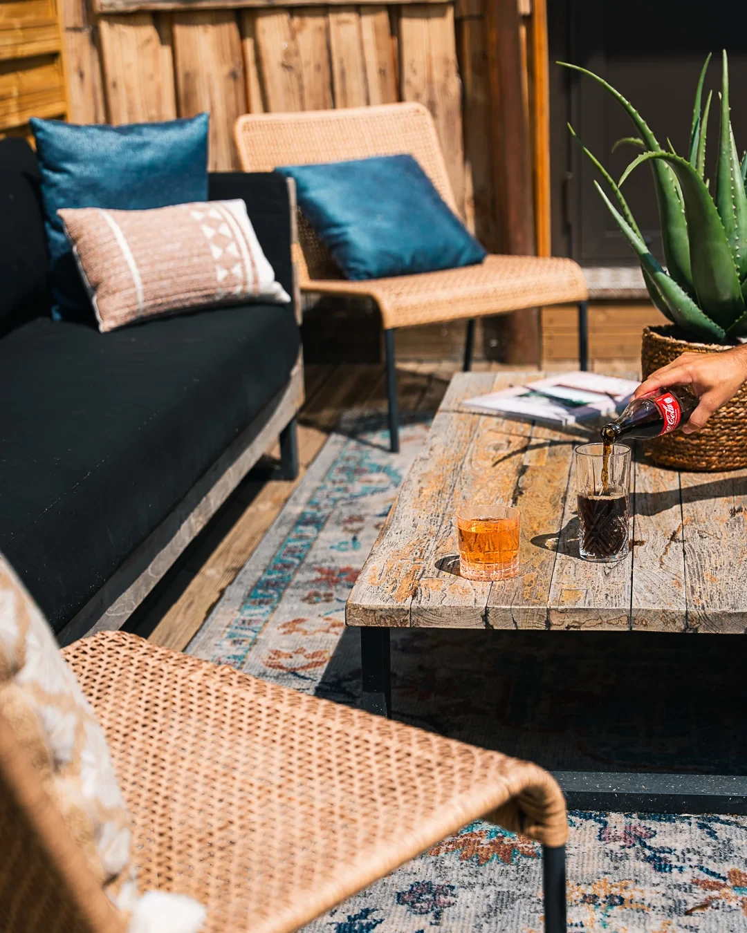Salon de terrasse de l’Hôtel La Chaudanne, avec fauteuils tressés, coussins et boissons sur une table en bois.