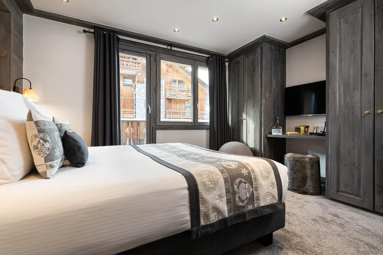 Chambre de l’Hôtel La Chaudanne avec lit double, boiseries grises et fenêtre donnant sur des chalets en bois.
