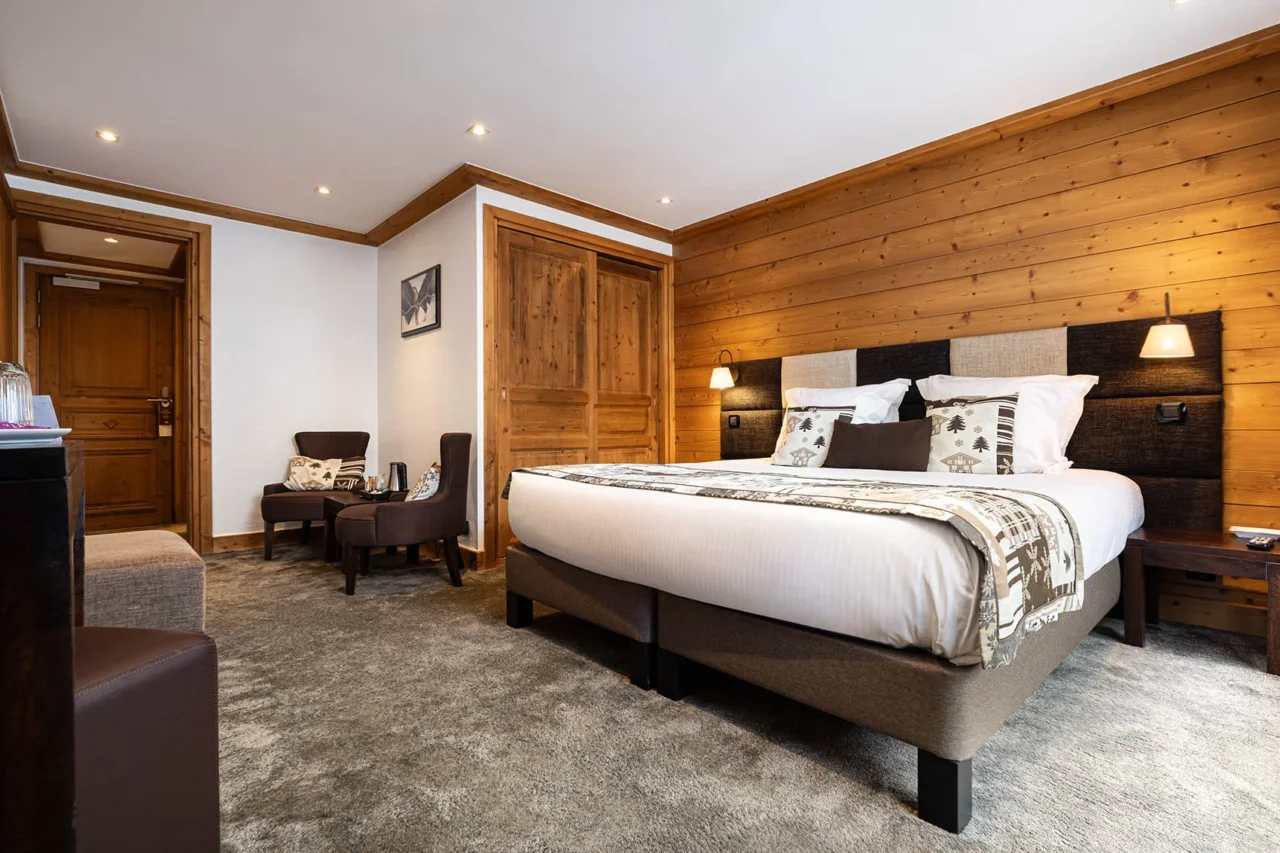 Chambre de l’Hôtel La Chaudanne, hôtel 4 étoiles à Méribel, avec lit double, mur en bois et coin salon à deux fauteuils.