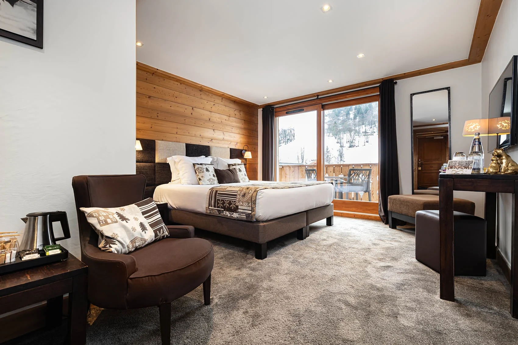 Chambre avec lit double et fauteuil, ouverte sur un balcon face à un versant boisé enneigé.